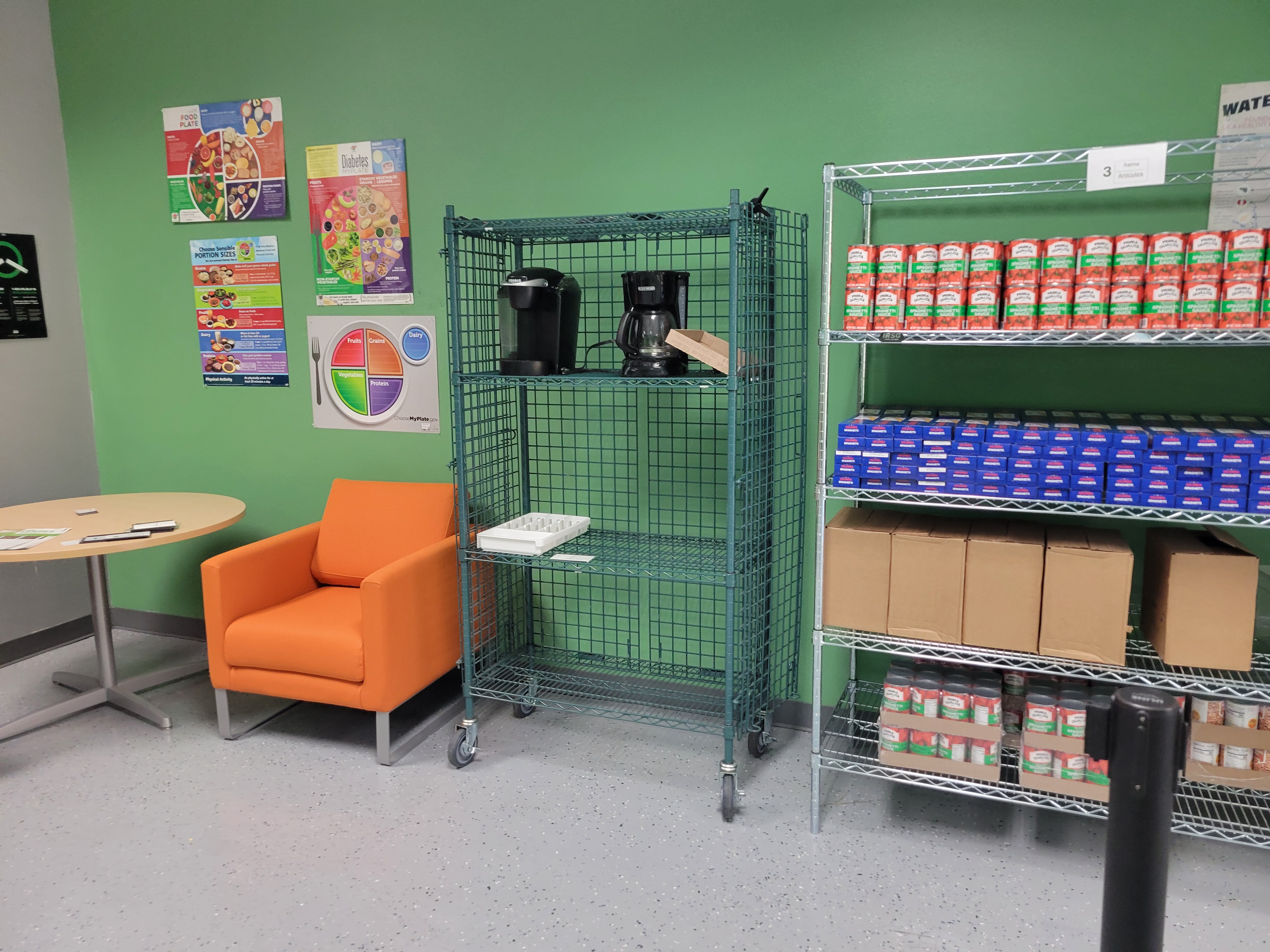 Table and chair next to cage shelf with two coffee markers on the top shelf next to food filled shelf.